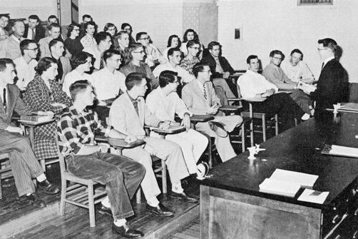 This black and white photo shows a professor on the left in front of a lecture hall with students seated at desks staggered on steps.